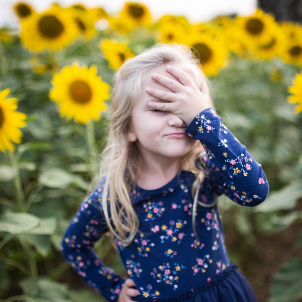 Das Bild zeigt ein Mädchen, das sich mit der linken Hand die Augen zuhält. Nach dem Motto: "Oh je, wie sieht das denn aus!". Im Hintergrund sind viele Sonnenblumen zu sehen.