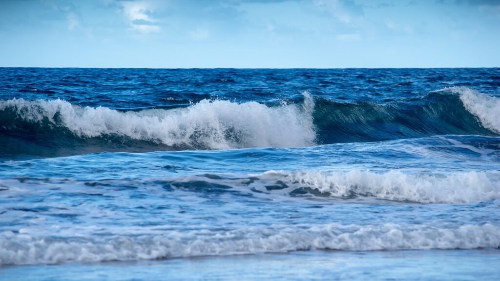 Das Bild zeigt blaues Meerwasser mit leichten Wellen und Schaumkronen. Oben ist blauer Himmel sichtbar.