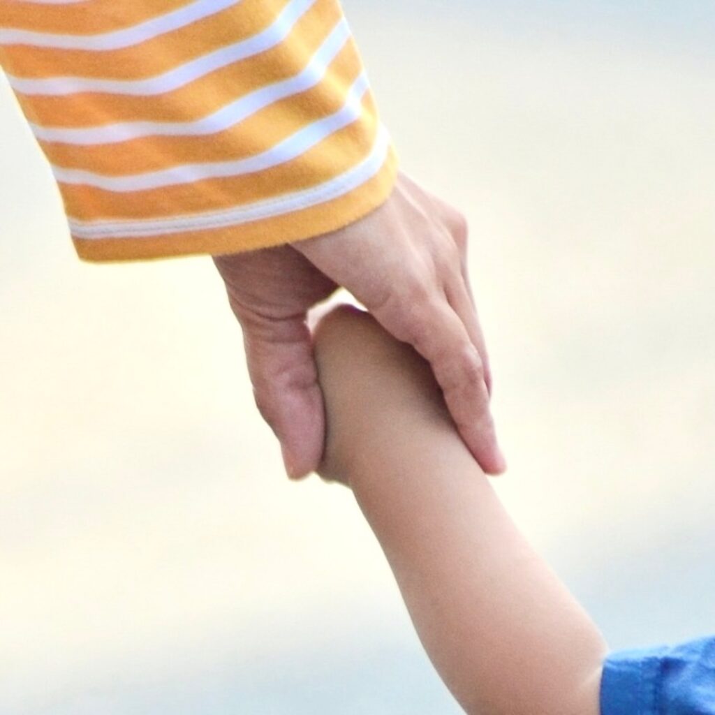 Das Bild zeigt einen nach unten geneigten Arm eines erwachsenen Menschen, mit einem gelb-weißen Pullover. Die Hand hält eine Kinderhand.