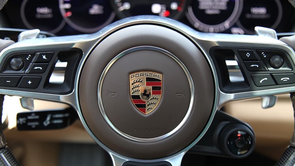 Das Bild zeigt das Lenkrad eines Porsche-Autos. In der Mitte das Porsche-Logo. Im Hintergrund Instrumente des Cockpits.